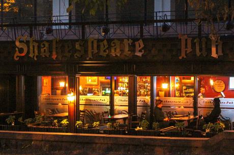 The illuminated sign of the Shakespeare Pub in Almaty glowing in the evening, with a few patrons visible inside through the large windows.