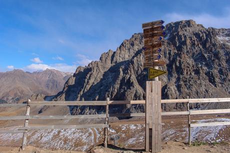 A wooden signpost with multiple directional arrows pointing to different locations around the world, set against a rugged mountain backdrop, at an altitude of 3,200 meters.