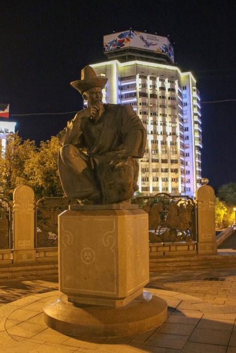 A bronze statue of a man in traditional attire, sitting thoughtfully with one hand on his chin, is illuminated against a city backdrop. The statue is placed on a decorated pedestal, with a brightly lit tall building in the background.