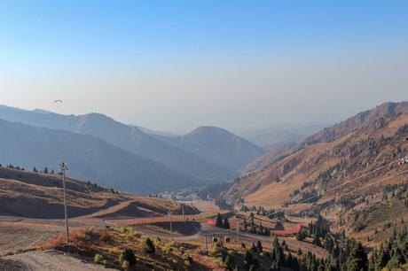 A paraglider in the distance soars above a mountain valley, surrounded by hazy hills and a clear blue sky.