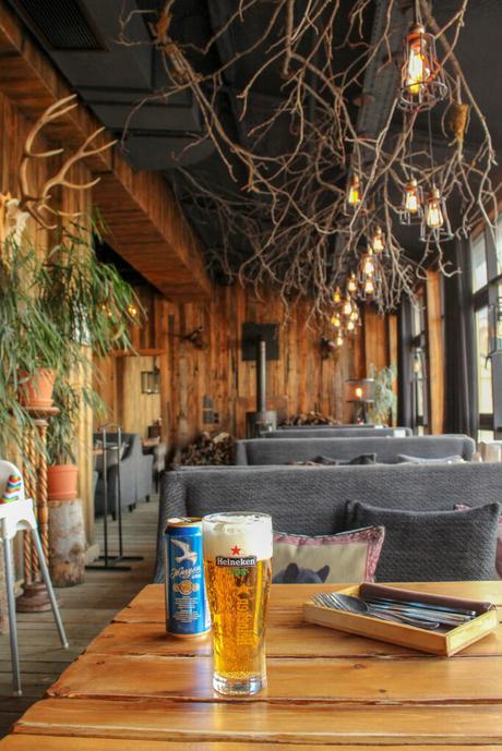 A cozy, rustic pub setting featuring wooden furniture, hanging branches as decor, and a Heineken beer and local brew placed on a wooden table.