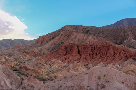 A landscape of vibrant red and yellow canyon walls, with rolling hills in the foreground and soft clouds overhead, showcasing the eroded textures of the cliffs.