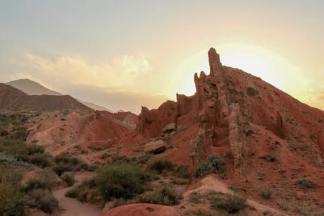 Sun setting behind jagged red rock formations in a desert-like landscape, casting a soft glow over the rugged terrain.