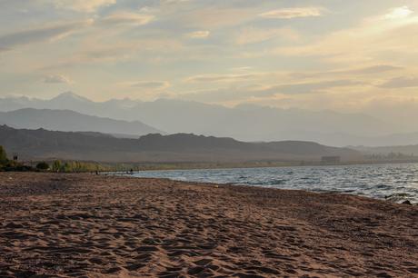 A peaceful lakeside view at sunset, with soft golden light reflecting off the water. The distant mountains create a layered backdrop, giving the image a sense of tranquility and natural beauty.