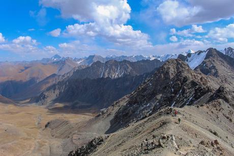 A panoramic scene of jagged mountain ridges and barren valleys under a vast blue sky, as hikers walk along a narrow trail on a rocky ridge, dwarfed by the dramatic landscape.