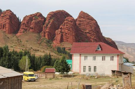 A white building with a red roof stands in front of towering red rock formations, with a yellow van parked nearby in a rural landscape.