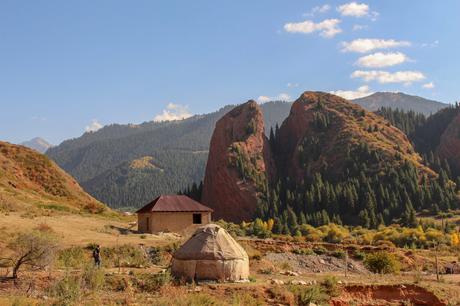 A traditional yurt sits in the foreground of a scenic valley, with towering red cliffs and lush green pine forests in the background. The earthy tones of the landscape highlight the natural beauty of the region.