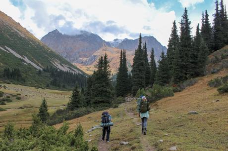 Two hikers with backpacks walk along a mountain path surrounded by pine trees and rugged peaks under cloudy skies.