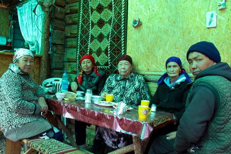 A group of older Kyrgyz people seated around a table inside a wooden house, enjoying a meal together. Their traditional headscarves and clothing add cultural depth to the cozy setting, while a woven tapestry decorates the wall.