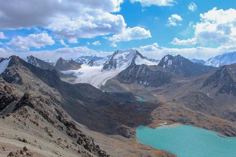 A sweeping view of a snow-covered glacier flowing between rocky mountain ridges, with multiple smaller lakes dotting the rugged terrain below. The peaks of distant mountains stand tall against a clear, cloud-dappled sky.