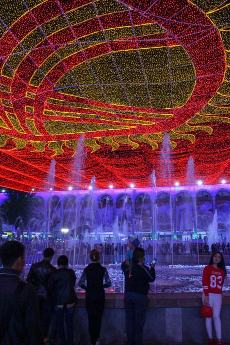 A fountain with people gathered around, illuminated by a large overhead display of bright red and yellow lights. The night scene feels lively and festive.