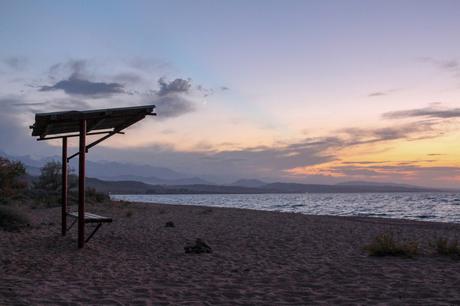 A quiet beach scene on Lake Issyk Kul at sunset, with a small wooden structure silhouetted against the evening sky. The calm waters of the lake and distant mountains are visible, while the sky is painted with hues of orange and purple.