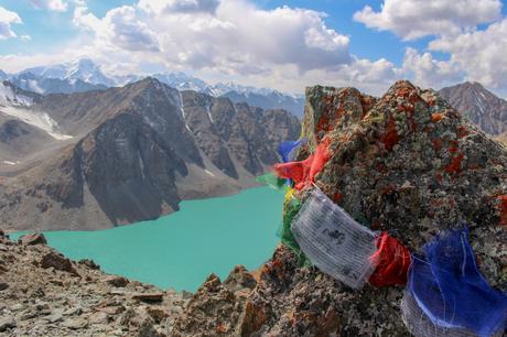 Colorful prayer flags tied to rocks flutter in the wind on a mountain summit overlooking a turquoise lake, with snowy peaks in the distance.