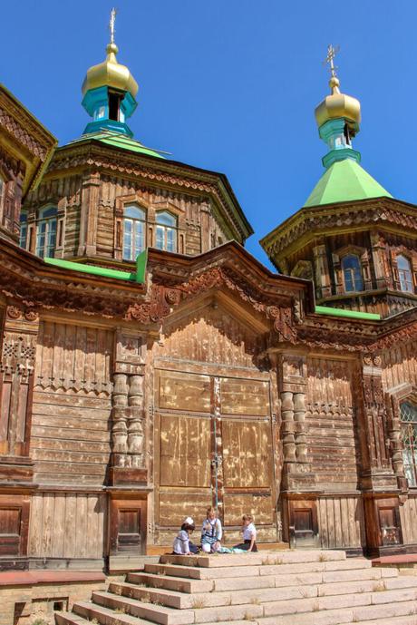 An old wooden church in the town of Karakol with ornate carvings and gold-domed towers under a bright blue sky. A group of people, including children, sit on the church's steps, adding a touch of human connection to the historical structure.