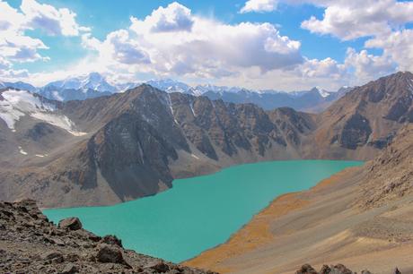 A stunning view of a glacial lake with turquoise waters surrounded by rugged mountains under a bright blue sky. Snow-capped peaks are visible in the distance, adding to the dramatic alpine landscape.