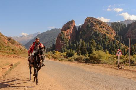 A local Kyrgyz man on horseback rides along a quiet road, with striking red rock formations and forested hills in the background. The serene landscape captures the essence of rural life.