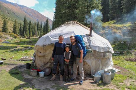 Alex Tiffany stands with a father and son outside their yurt in a mountainous valley, surrounded by trees and hills.