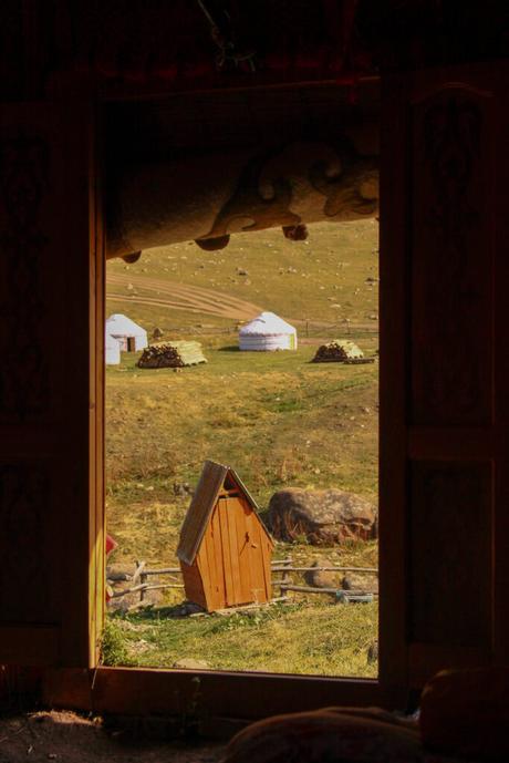 The view from the inside of a traditional Central Asian yurt, looking out onto a grassy steppe where more yurts are pitched. The landscape is framed by the doorway, with an outhouse and woodpile in the foreground.