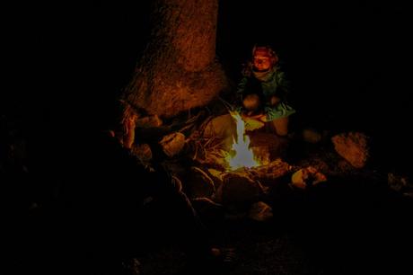 Two people sit around a small campfire at night, their faces softly illuminated by the flames, with a large tree trunk visible behind them.