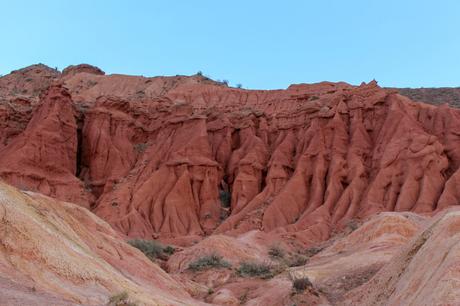 Close-up of striking red rock cliffs with sharp ridges and weathered textures, framed against a pale blue sky.