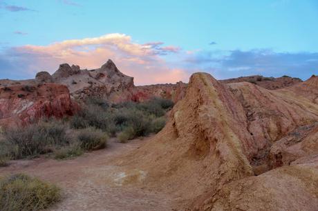 A trail through desert terrain with multicolored rock formations, ranging from red to beige, under a pastel sky with pink and blue clouds.