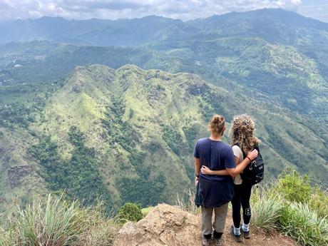 Two people standing at the edge of a cliff, embracing each other, and looking out over a mountainous valley covered in greenery.