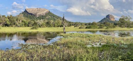 Two large rocky outcrops - Sigiriya Rock and Pidurangala - standing amidst green fields and a serene water body, reflecting the blue sky and clouds.