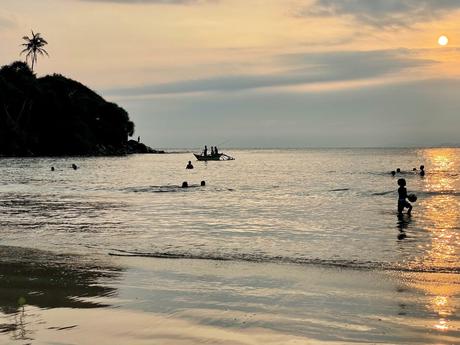 People swimming in the sea at sunset, with the golden light reflecting off the water and a small boat silhouetted against the horizon.