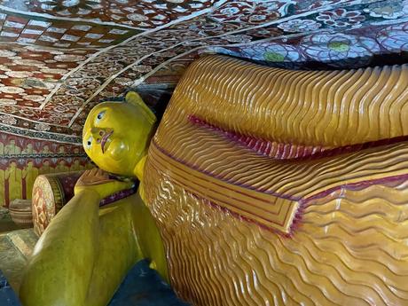 A large reclining Buddha statue inside a cave temple, surrounded by intricately painted walls and ceiling.