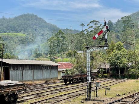 An old-fashioned railway signal post next to train tracks with a scenic backdrop of hills and greenery under a clear blue sky.