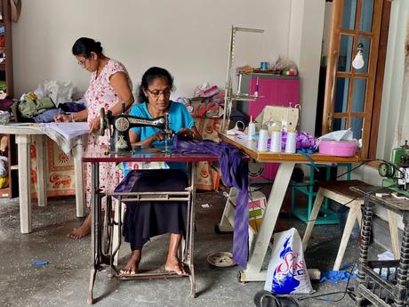 Two women working in a small sewing workshop, one sewing fabric on an old-fashioned sewing machine while the other cuts and measures fabric.