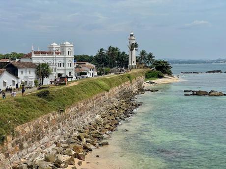 Galle Fort’s iconic lighthouse standing tall by the sea, surrounded by historic white buildings and a stone wall along the coastline.