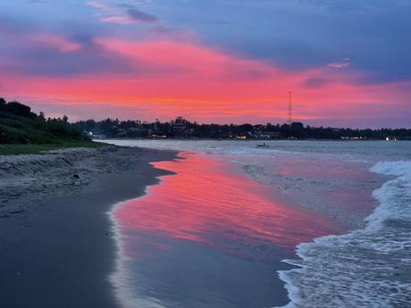 A stunning sunset on a beach, with vibrant hues of pink, purple, and orange reflecting off the water, and a serene, darkened shoreline.