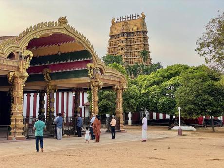 A large temple complex with intricate golden architecture, surrounded by people observing and walking on the sandy grounds.