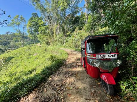 A red tuk-tuk parked on a dirt road surrounded by lush green hills and trees in a rural area in central Sri Lanka.