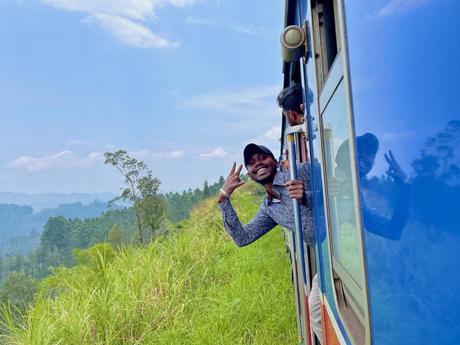 A man leaning out of a train window with a big smile, giving a peace sign with his hand while the train moves through a lush green countryside.