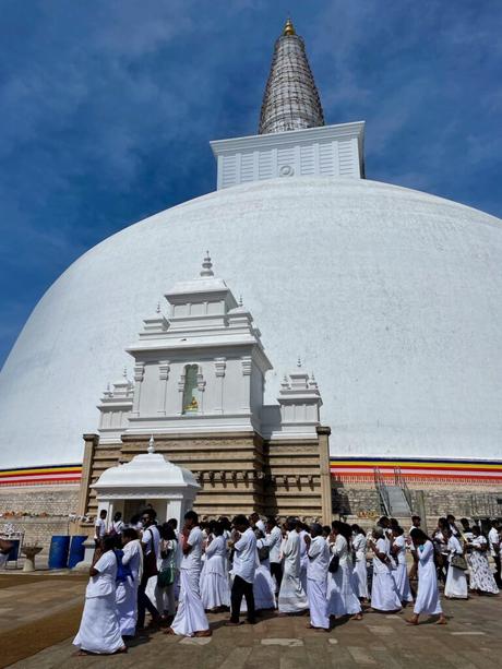 A large white stupa with a golden top, surrounded by worshippers dressed in white, walking in a ceremonial procession around the structure.