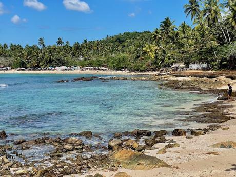 The calm beach at Hiriketiya with clear turquoise water, surrounded by lush palm trees and rocky outcrops.