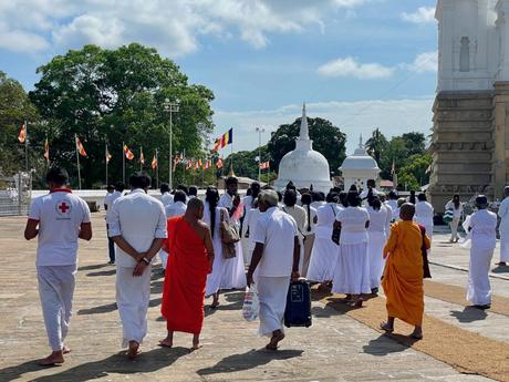 A procession of worshippers, including Buddhist monks, walking barefoot in a temple courtyard, with white stupas and flags visible in the background.