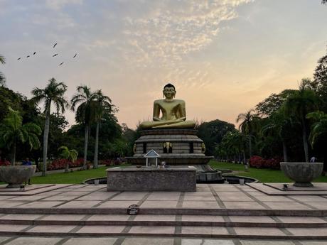 A large golden Buddha statue sits in a peaceful meditation posture on a stone platform in a landscaped garden. Palm trees and neatly trimmed bushes line the path leading up to the statue, with a group of birds flying in the sky during sunset, adding to the serene ambiance.