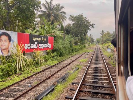 View from a train window of a railroad track with a large red billboard featuring a politician's face and text in Sinhala, surrounded by tropical vegetation.