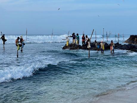 A group of traditional Sri Lankan stilt fishermen sitting on wooden poles in the ocean, casting their fishing lines into the waves. The blue water crashes around them as they work against the horizon.