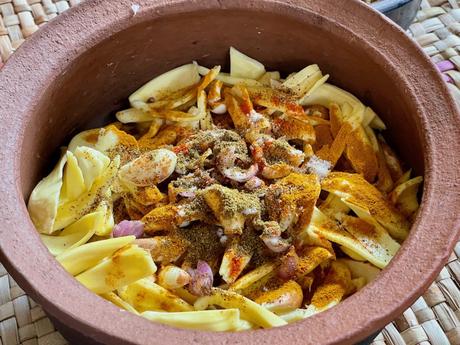 A clay pot filled with sliced jackfruit, onions, and various spices like turmeric and chili, preparing for a traditional Sri Lankan dish.