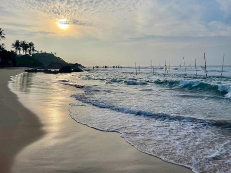 A beach at sunrise with calm waves reflecting the early morning light. In the distance, traditional stilt fishermen are perched on poles, creating a peaceful coastal scene.