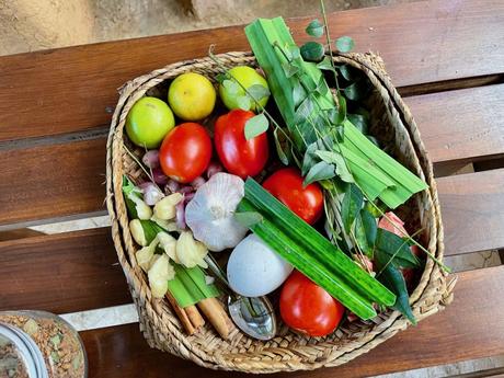 A basket filled with fresh ingredients including tomatoes, garlic, curry leaves, limes, and other vegetables, placed on a wooden surface ready for cooking.