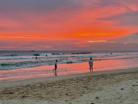 A vibrant sunset reflecting on the ocean with a few surfers and beachgoers standing at the water's edge, enjoying the serene view.