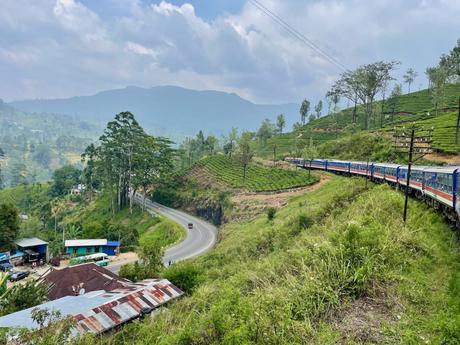 A long passenger train winding through hilly tea plantations and curving roads in the Sri Lankan highlands, with mountains in the background.