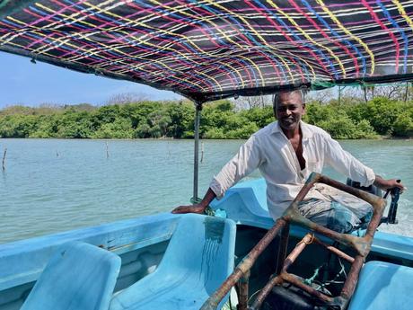 A man smiling while steering a small blue boat along a calm river, with a colorful roof overhead and dense greenery in the background.