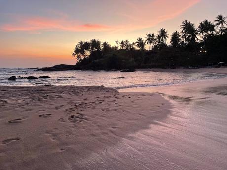 A serene sunset over a beach, with pink hues in the sky and footprints in the sand leading towards the water.