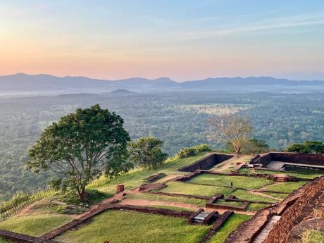 A panoramic view of green fields and ruins from an elevated viewpoint, with mountains stretching across the horizon at sunset.